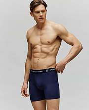 A muscular man stands against a plain background wearing Psycho Bunny MEN'S 3 PACK BOXER BRIEF (MO1400006) in navy blue, featuring a black waistband with “Psycho Bunny” in white text. He looks to the side, one hand touching the waistband.