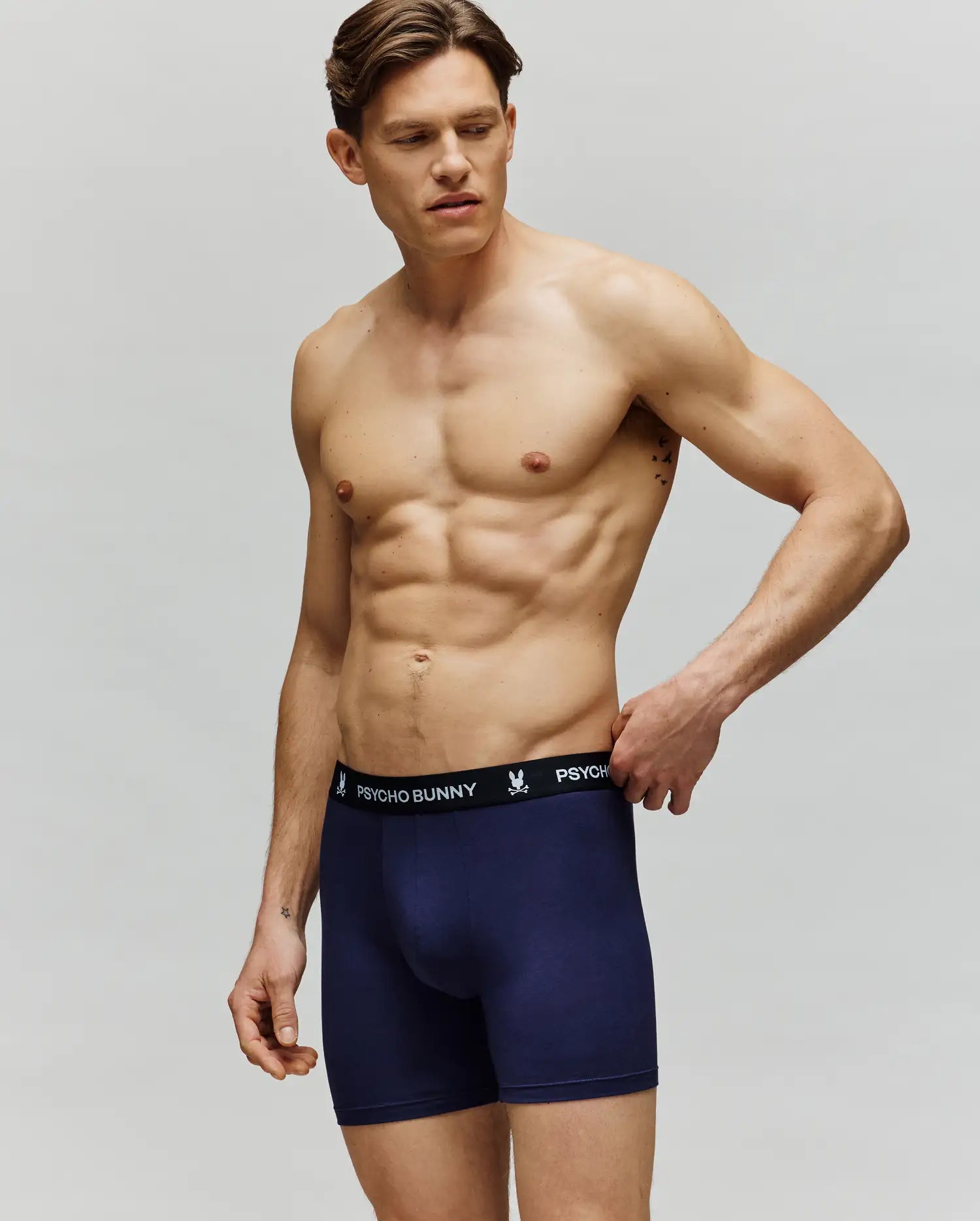 A muscular man stands against a plain background wearing Psycho Bunny MEN'S 3 PACK BOXER BRIEF (MO1400006) in navy blue, featuring a black waistband with “Psycho Bunny” in white text. He looks to the side, one hand touching the waistband.