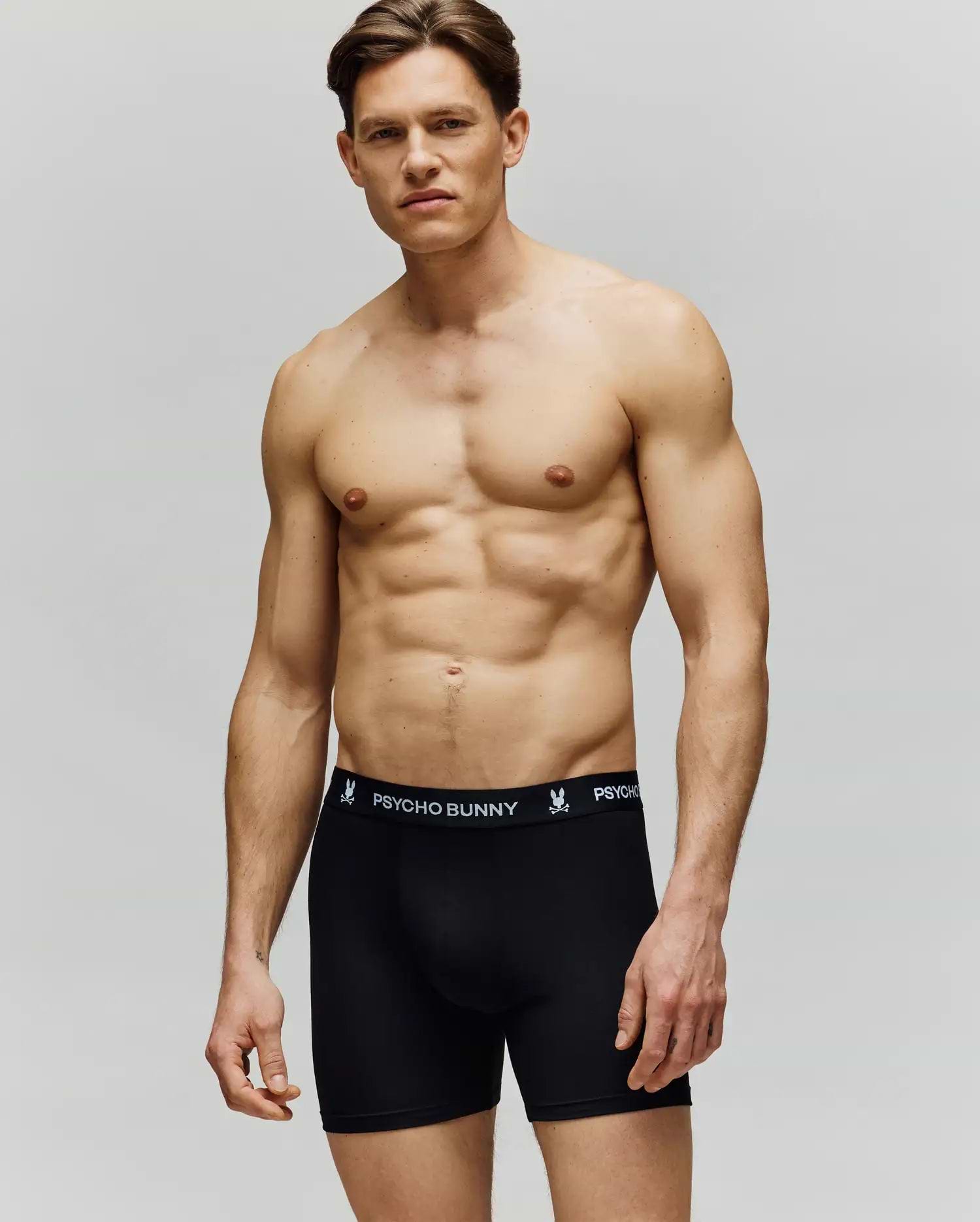 A fit young man with short brown hair stands shirtless against a plain background, wearing Psycho Bunny's MENS 3 PACK BOXER BRIEF - MO1400006 in black, featuring the brand logo on the waistband.