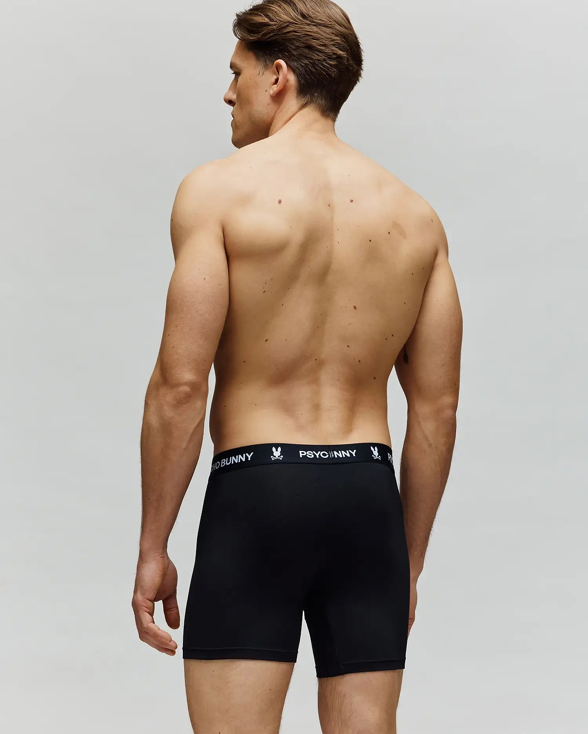 A man faces away, shirtless, wearing Psycho Bunny MENS 3 PACK BOXER BRIEF - MO1400006. The black cotton-modal briefs have a branded waistband with a bunny logo. The background is plain and light-colored.