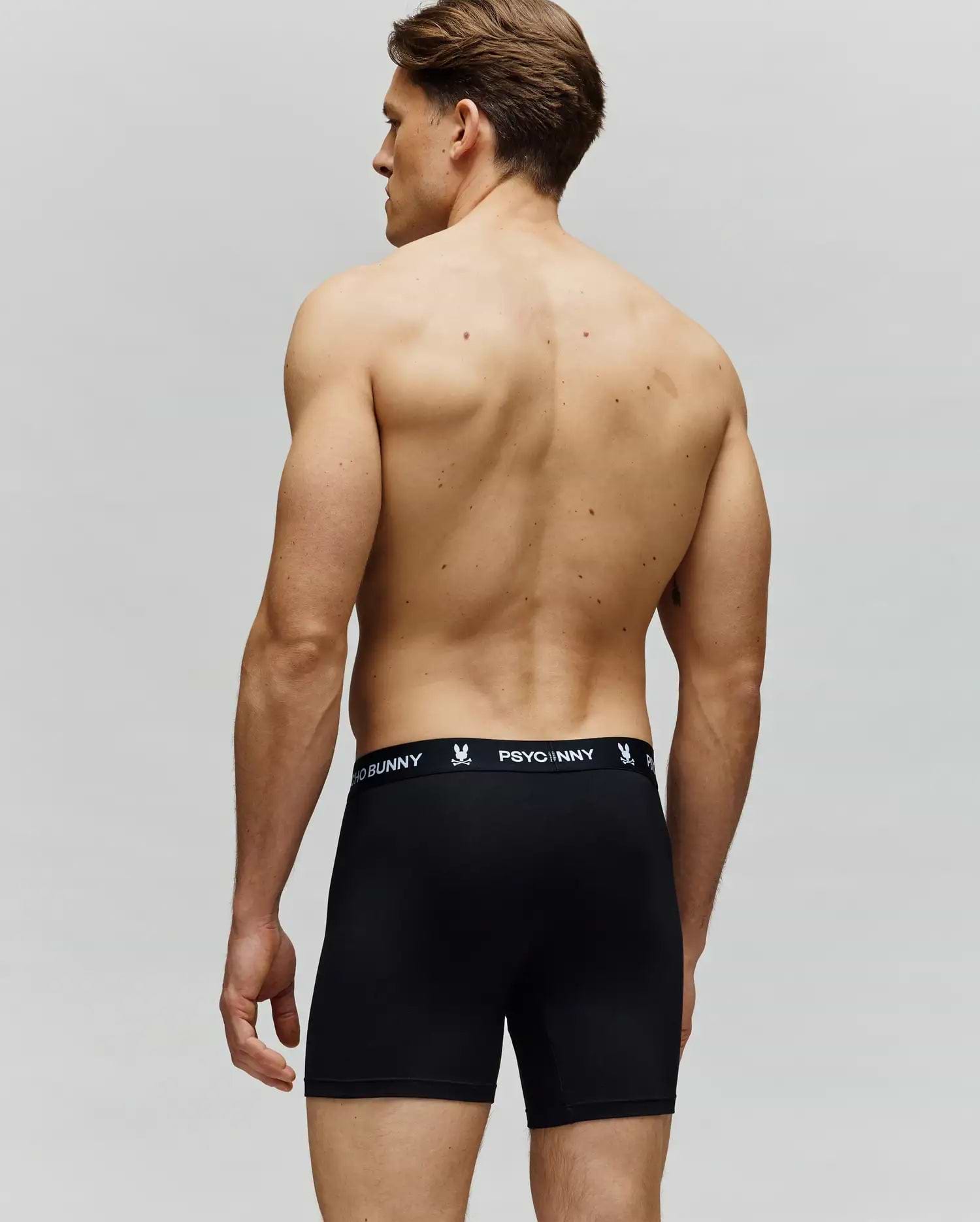 A shirtless man faces away, wearing Psycho Bunny MENS 3 PACK BOXER BRIEF - MO1400006 in black with a branded waistband. Made from a cotton-modal blend, these boxer briefs deliver all-day comfort against a neutral background.