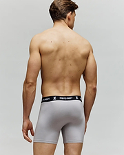 A man stands with his back to the camera, wearing Psycho Bunny MENS 3 PACK BOXER BRIEF - MO1400006 in light gray with a black "PSYCHO BUNNY" waistband, set against a plain neutral background.