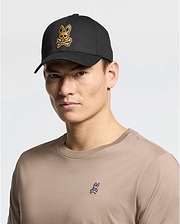 A person wears a beige 100% cotton t-shirt with a small rabbit logo, paired with the Psycho Bunny MENS LAS VEGAS BASEBALL CAP in black, featuring a direct embroidery yellow skull and crossbones rabbit logo. The background is plain white.