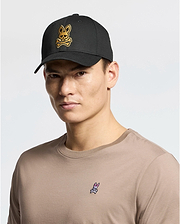 A person wears a beige 100% cotton t-shirt with a small rabbit logo, paired with the Psycho Bunny MENS LAS VEGAS BASEBALL CAP in black, featuring a direct embroidery yellow skull and crossbones rabbit logo. The background is plain white.