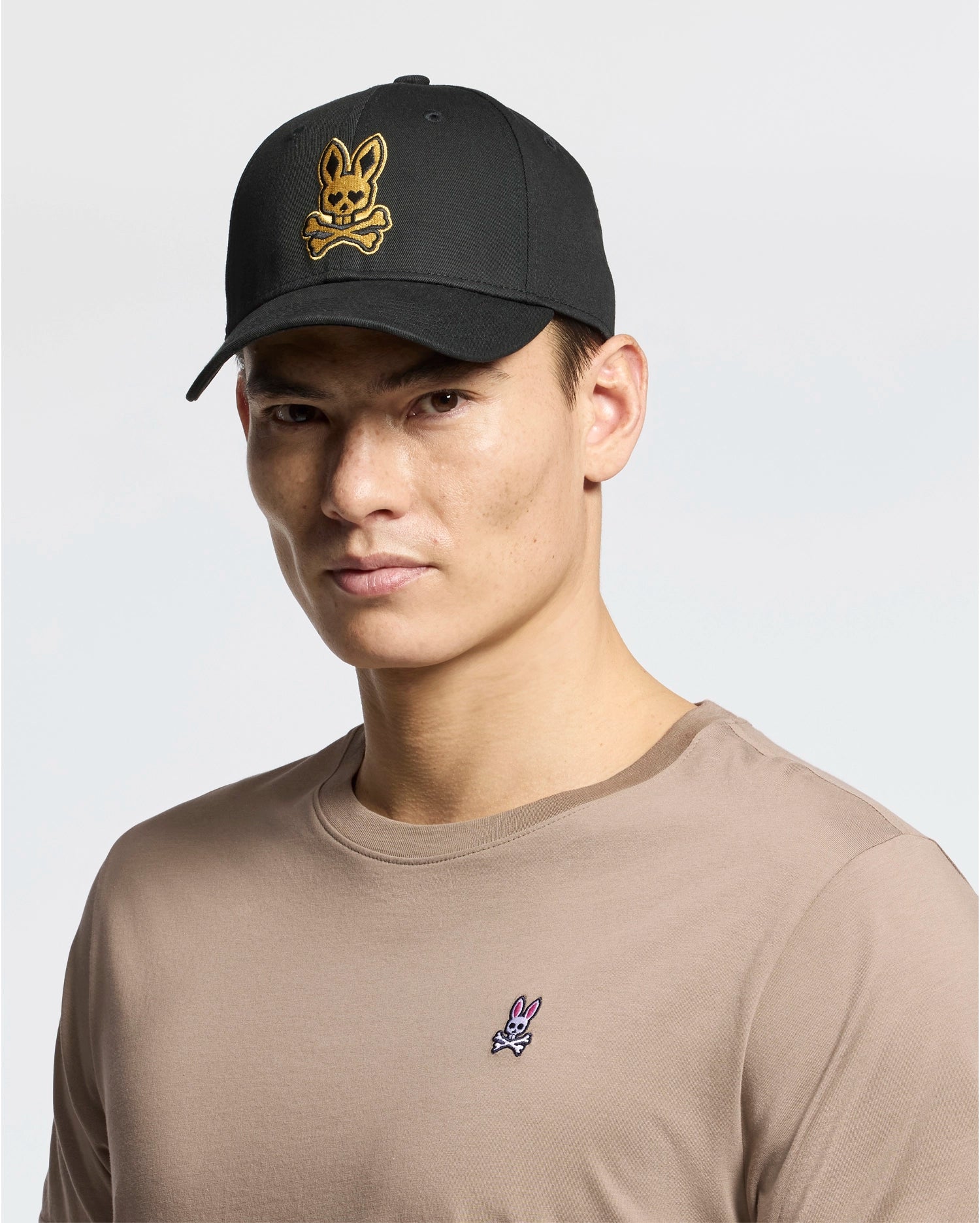A person wears a beige 100% cotton t-shirt with a small rabbit logo, paired with the Psycho Bunny MENS LAS VEGAS BASEBALL CAP in black, featuring a direct embroidery yellow skull and crossbones rabbit logo. The background is plain white.