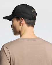 Seen from behind, a person wears the MENS LAS VEGAS BASEBALL CAP - MO1500011 by Psycho Bunny, featuring "LAS VEGAS" in yellow direct embroidery on black. Their short dark hair contrasts with a beige 100% cotton T-shirt against a plain, light background.