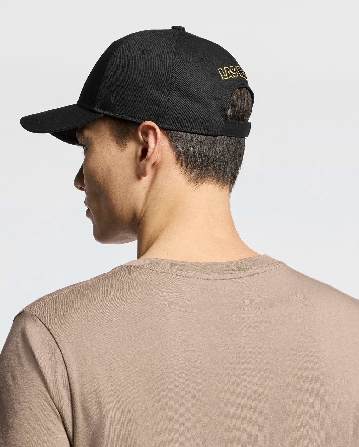 Seen from behind, a person wears the MENS LAS VEGAS BASEBALL CAP - MO1500011 by Psycho Bunny, featuring "LAS VEGAS" in yellow direct embroidery on black. Their short dark hair contrasts with a beige 100% cotton T-shirt against a plain, light background.