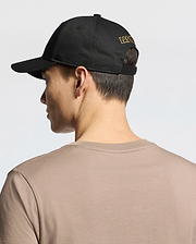 Seen from behind, a person wears the MENS LAS VEGAS BASEBALL CAP - MO1500011 by Psycho Bunny, featuring "LAS VEGAS" in yellow direct embroidery on black. Their short dark hair contrasts with a beige 100% cotton T-shirt against a plain, light background.