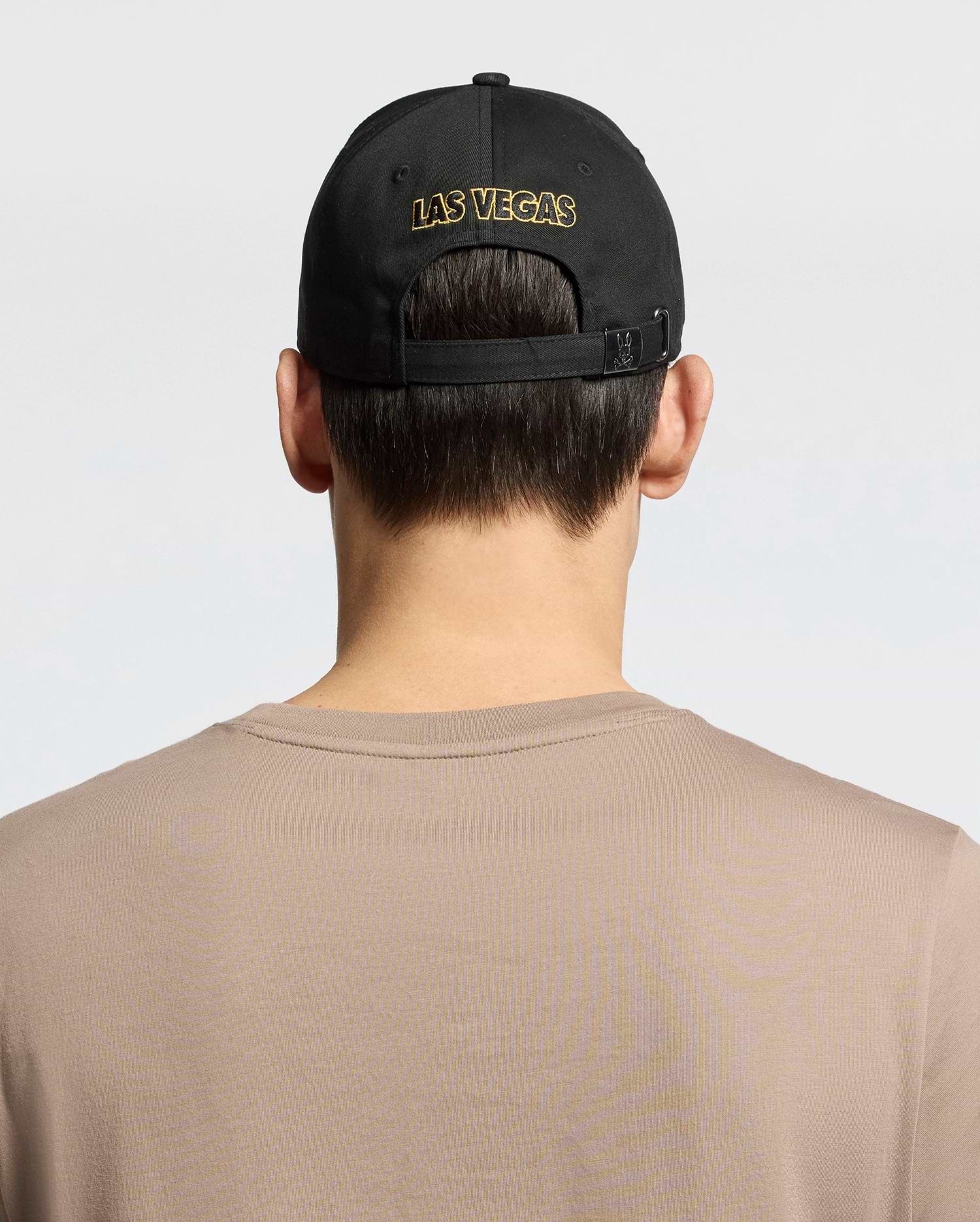 A person with short brown hair, wearing a Psycho Bunny MENS LAS VEGAS BASEBALL CAP - MO1500011 in black with "LAS VEGAS" stitched in gold via direct embroidery and a 100% cotton beige shirt, is shown from behind against a plain white backdrop.