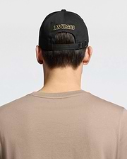 A person with short brown hair, wearing a Psycho Bunny MENS LAS VEGAS BASEBALL CAP - MO1500011 in black with "LAS VEGAS" stitched in gold via direct embroidery and a 100% cotton beige shirt, is shown from behind against a plain white backdrop.