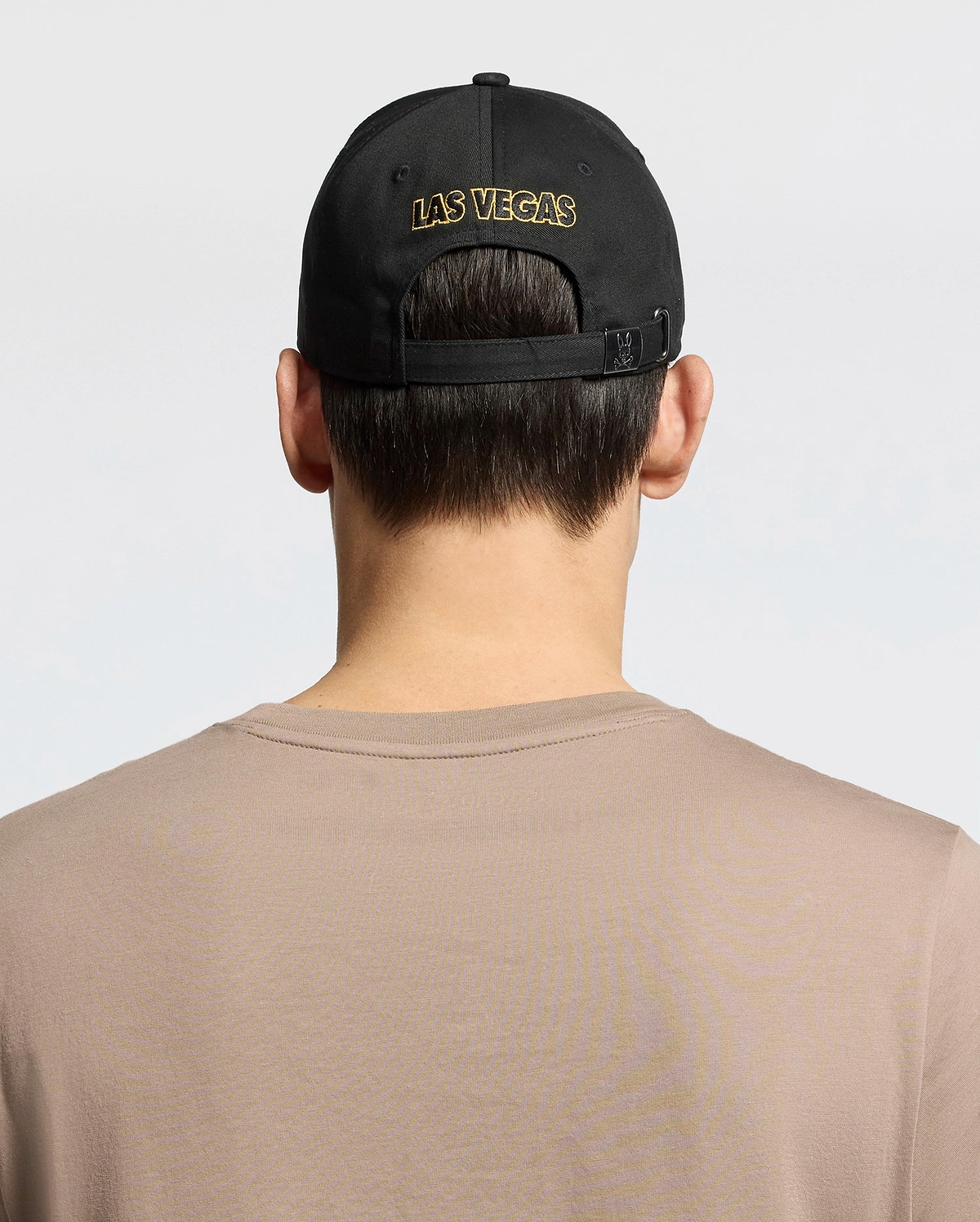 A person with short brown hair, wearing a Psycho Bunny MENS LAS VEGAS BASEBALL CAP - MO1500011 in black with "LAS VEGAS" stitched in gold via direct embroidery and a 100% cotton beige shirt, is shown from behind against a plain white backdrop.