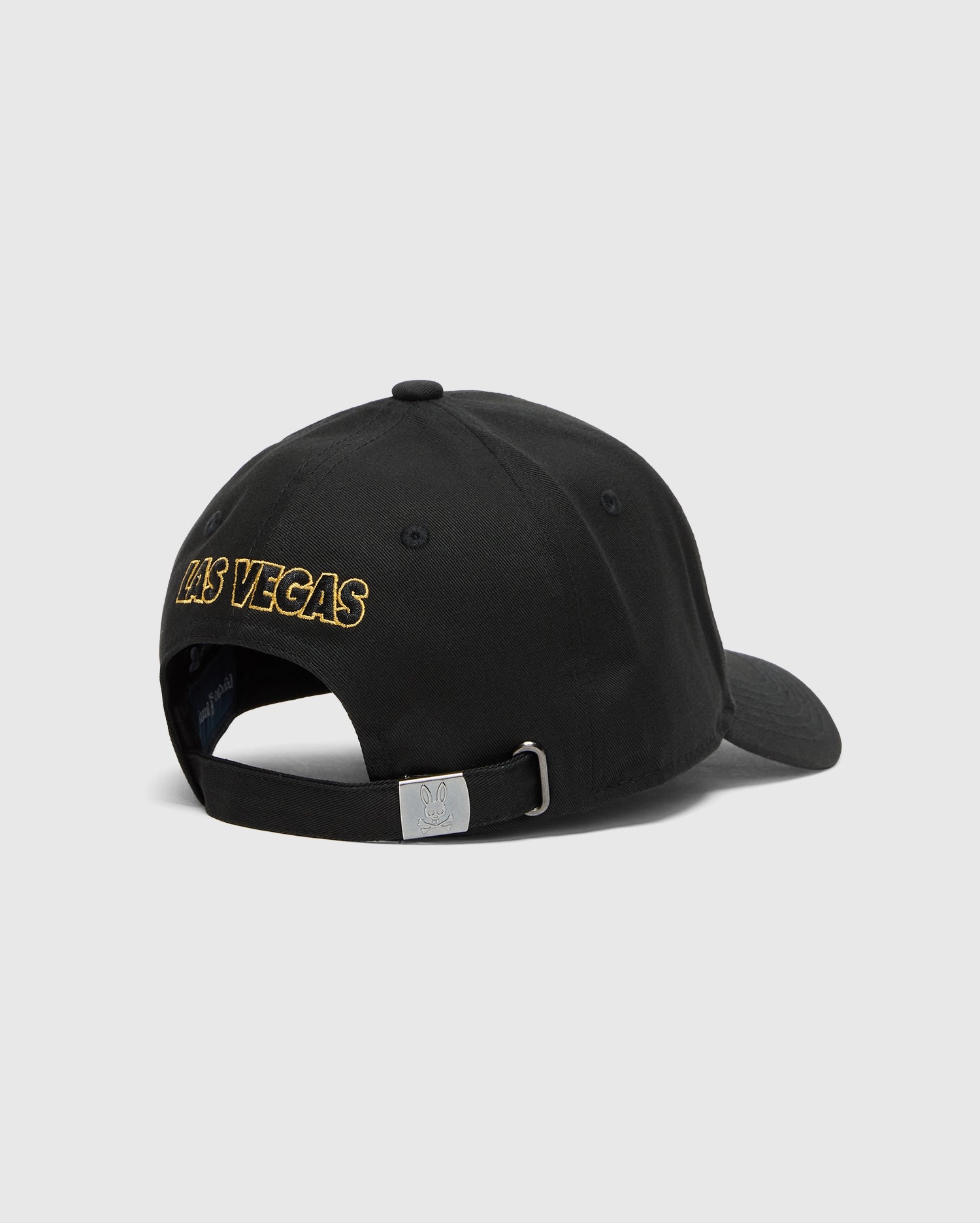 The Psycho Bunny MENS LAS VEGAS BASEBALL CAP (MO1500011) is a black, 100% cotton hat with gold "Las Vegas" embroidery on the back, direct embroidery, and an adjustable strap with a metal buckle. Made in China, it stands out beautifully against a plain white background.