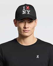 A man wears the Psycho Bunny MENS NEW YORK BASEBALL CAP - MO1500012, a black cotton cap with "I NY" and red embroidered bunny logo, plus a black shirt featuring a small pink and white embroidered bunny chest logo, against a light background.