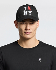 A man wears the Psycho Bunny MENS NEW YORK BASEBALL CAP - MO1500012, a black cotton cap with "I NY" and red embroidered bunny logo, plus a black shirt featuring a small pink and white embroidered bunny chest logo, against a light background.