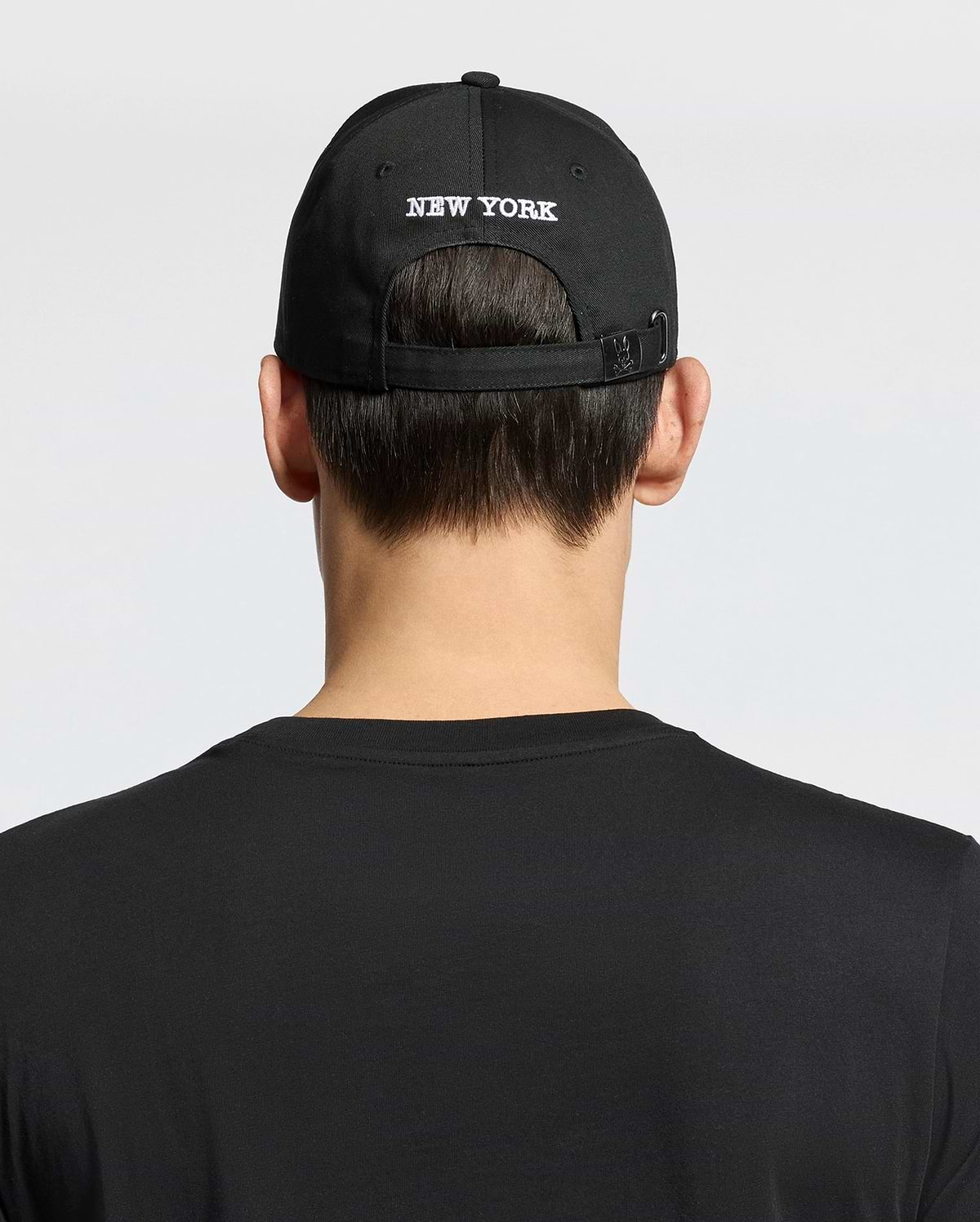 A person shown from behind against a plain background wears a woven black cotton T-shirt and the Psycho Bunny MENS NEW YORK BASEBALL CAP - MO1500012, featuring "NEW YORK" embroidered on the back.