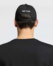A person shown from behind against a plain background wears a woven black cotton T-shirt and the Psycho Bunny MENS NEW YORK BASEBALL CAP - MO1500012, featuring "NEW YORK" embroidered on the back.