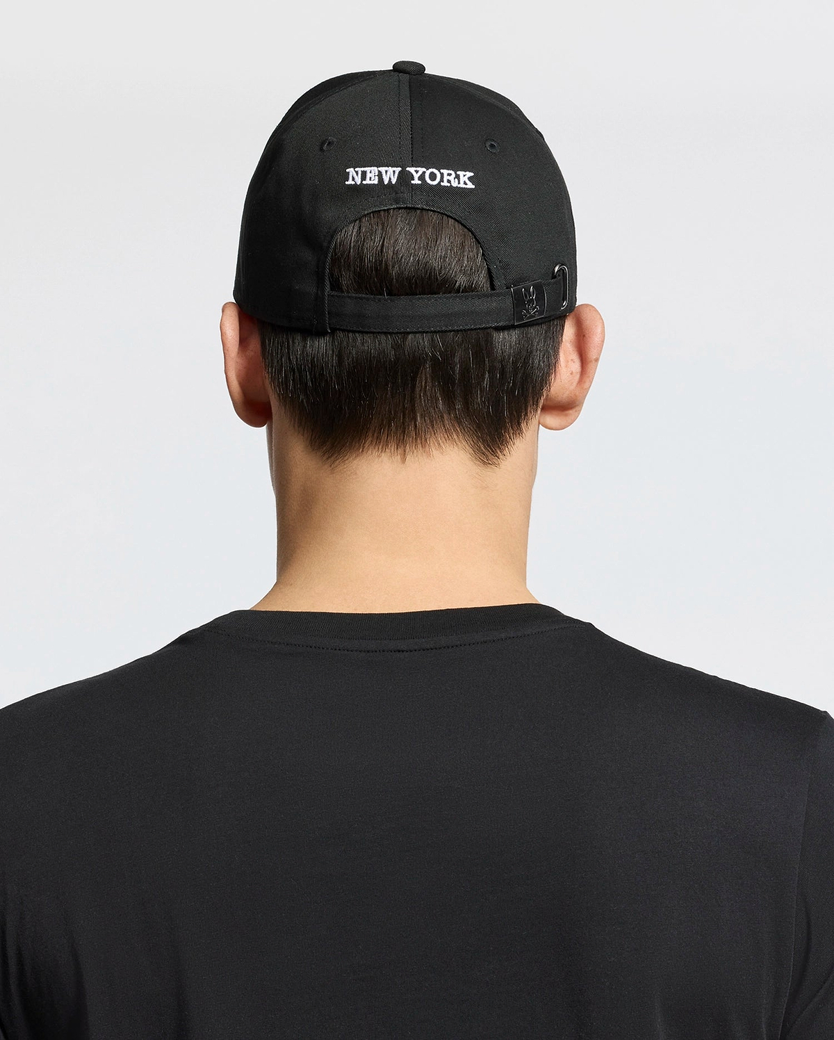 A person shown from behind against a plain background wears a woven black cotton T-shirt and the Psycho Bunny MENS NEW YORK BASEBALL CAP - MO1500012, featuring "NEW YORK" embroidered on the back.