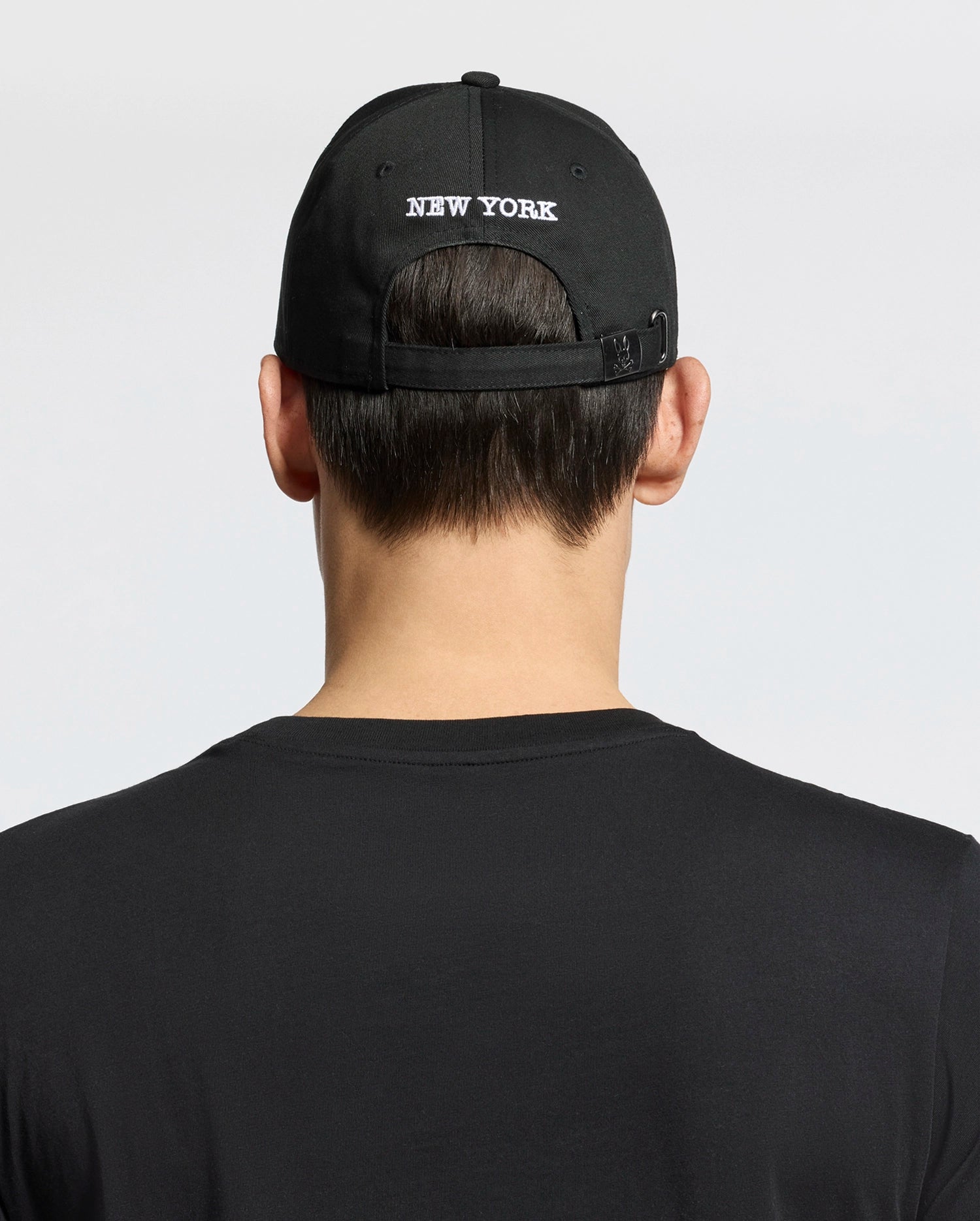 A person shown from behind against a plain background wears a woven black cotton T-shirt and the Psycho Bunny MENS NEW YORK BASEBALL CAP - MO1500012, featuring "NEW YORK" embroidered on the back.