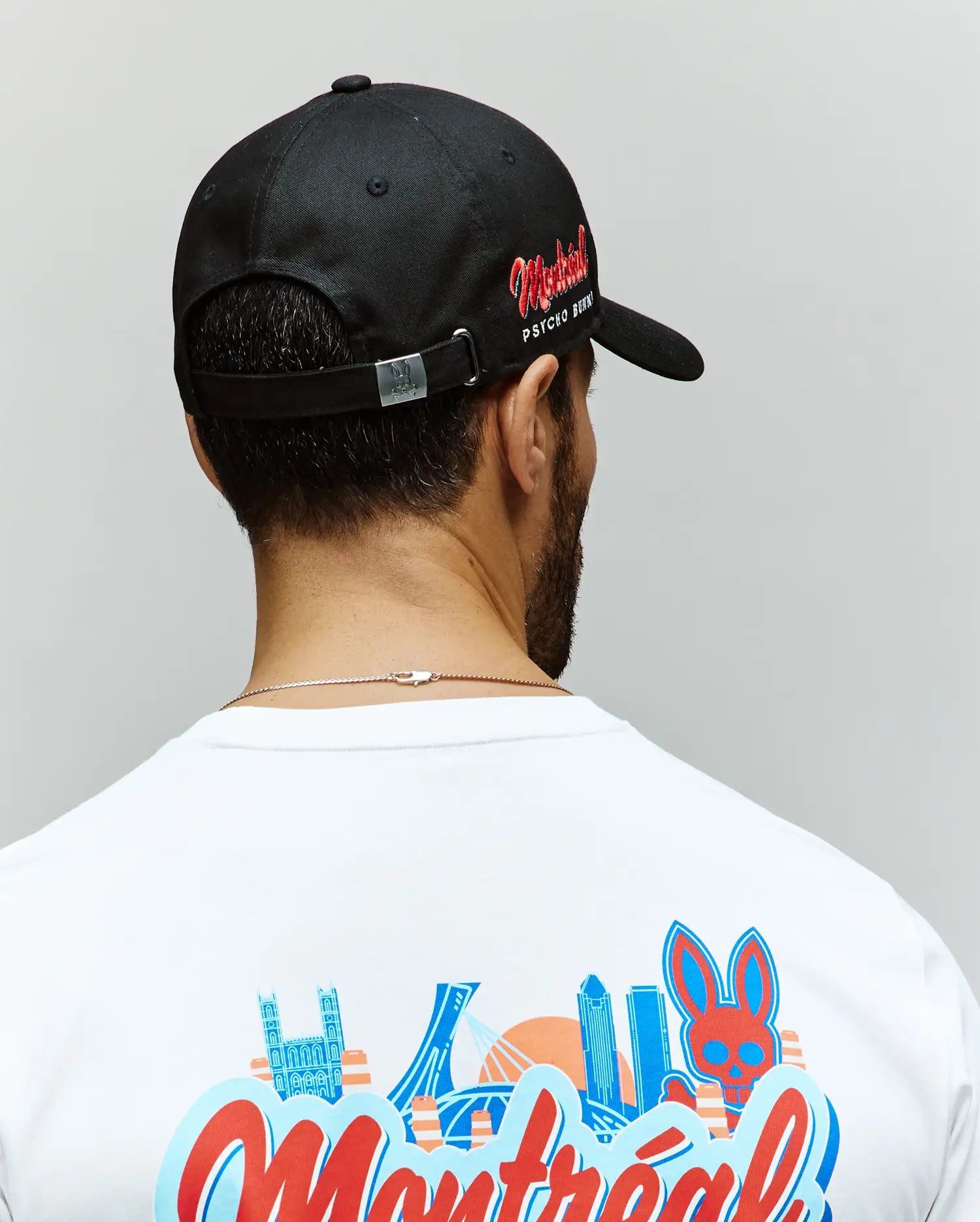 A person wears a black Psycho Bunny MENS MONTREAL BASEBALL CAP (MO1500048) and a white T-shirt with colorful Montreal graphics on the back, standing with their back to the camera against a plain light background.