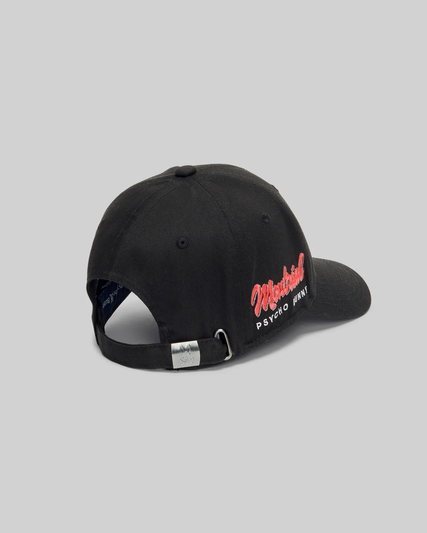 The Psycho Bunny MENS MONTREAL BASEBALL CAP (MO1500048) is a black 100% cotton cap with "Montreal PSYCHO PRINT" embroidered in red and white on the back left, plus an adjustable strap and metal buckle, shown on a light gray background.