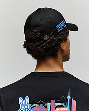A person with curly hair, seen from behind, wears the Psycho Bunny MENS TORONTO BASEBALL CAP (MO1500049) featuring blue "TORONTO PSYCHO BUNNY" text and an embroidered shirt with colorful Toronto landmarks.
