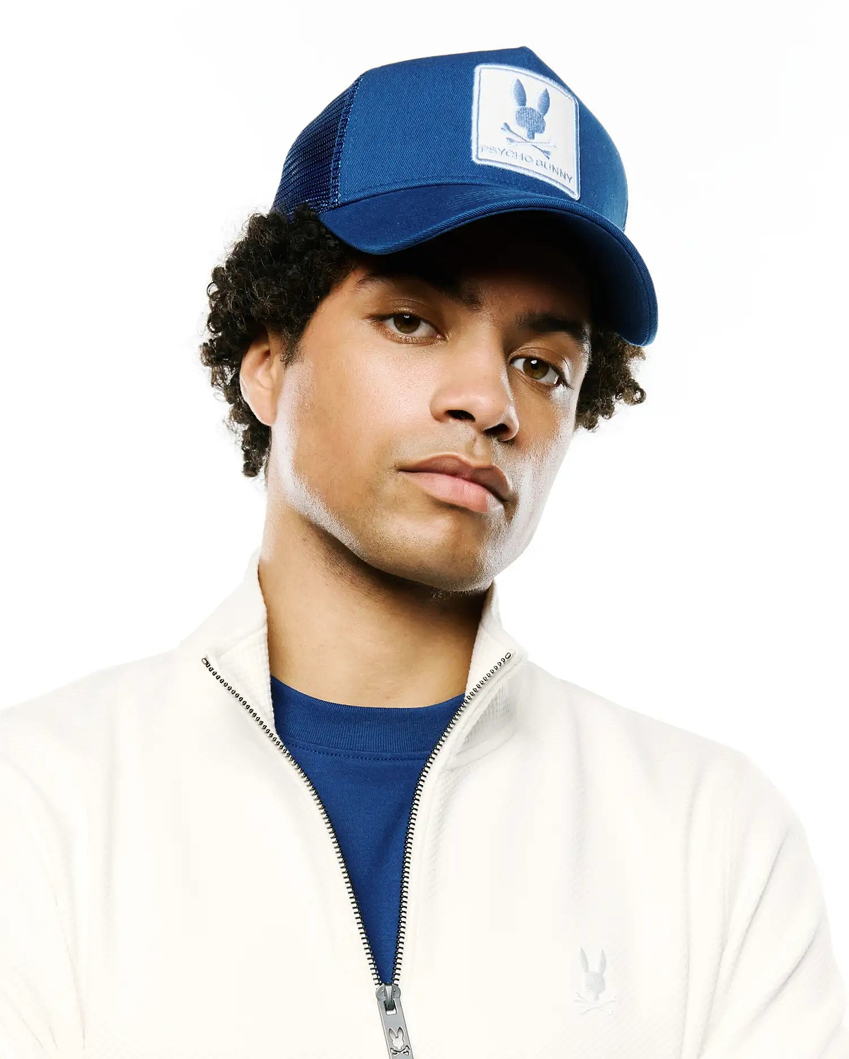 MENS ADAMO BUNNY TRUCKER CAP in royal blue with a white embroidered Psycho Bunny skull logo patch, an off-white textured full-zip jacket with a tonal chest logo, and a blue jersey crewneck t-shirt.