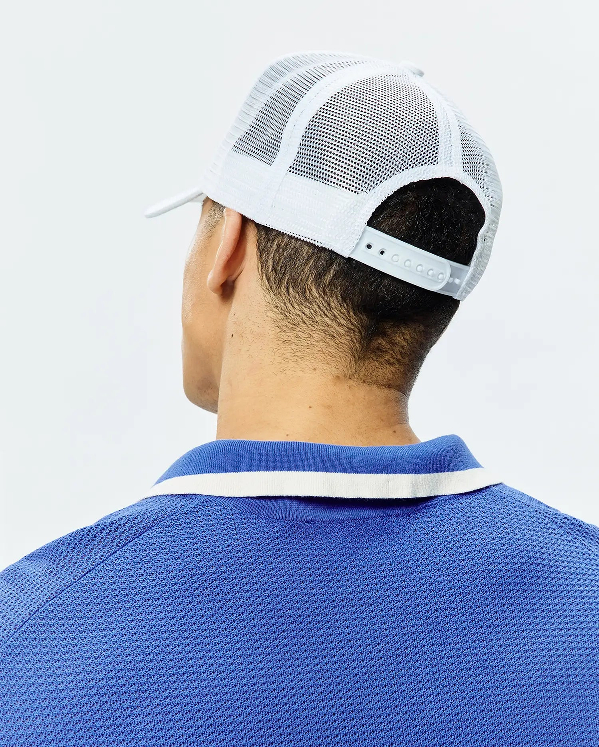 White Mens Adamo Trucker Cap with mesh back and snap closure, paired with a royal blue pique polo shirt featuring a blue and white striped collar.