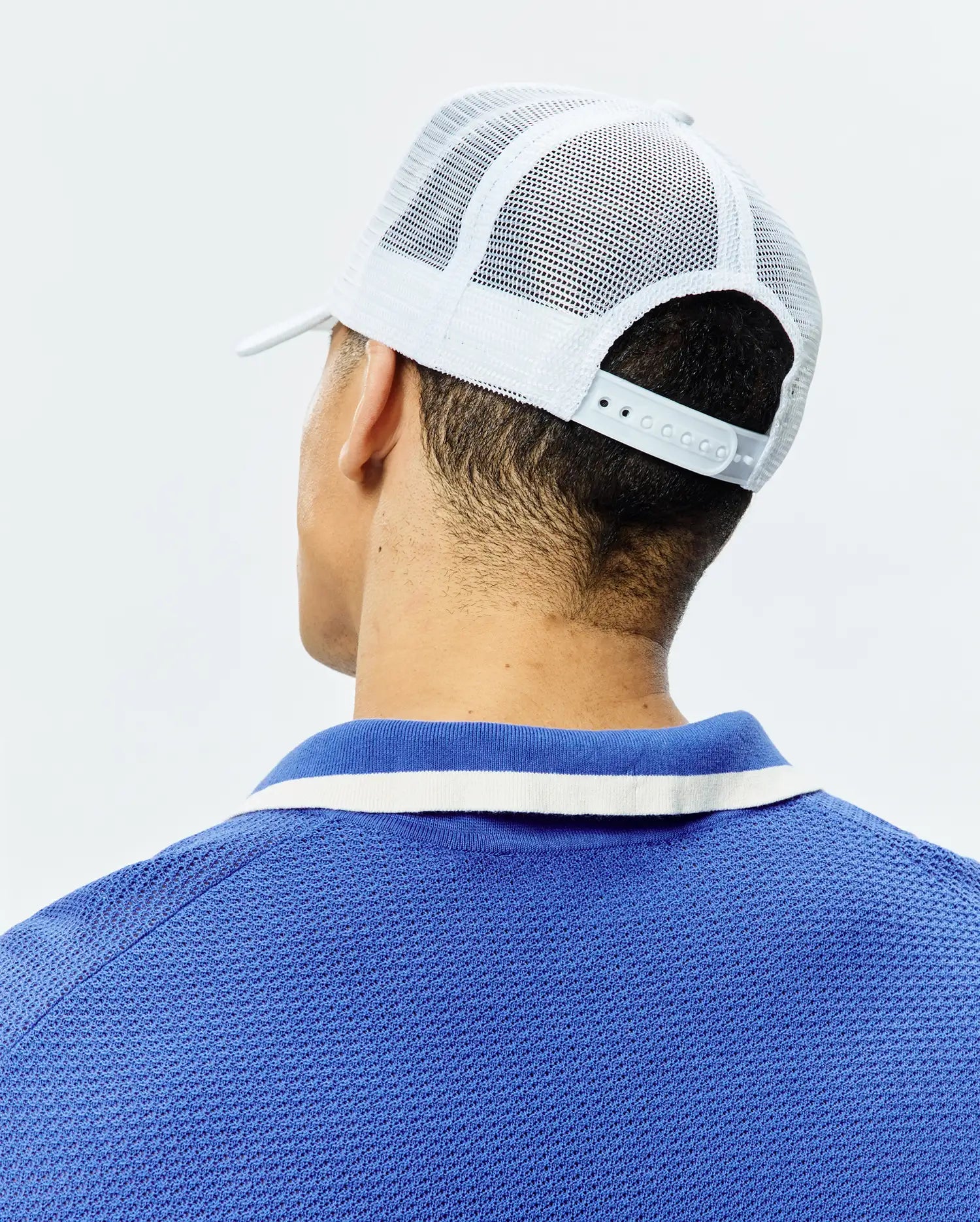 White Mens Adamo Trucker Cap with mesh back and snap closure, paired with a royal blue pique polo shirt featuring a blue and white striped collar.