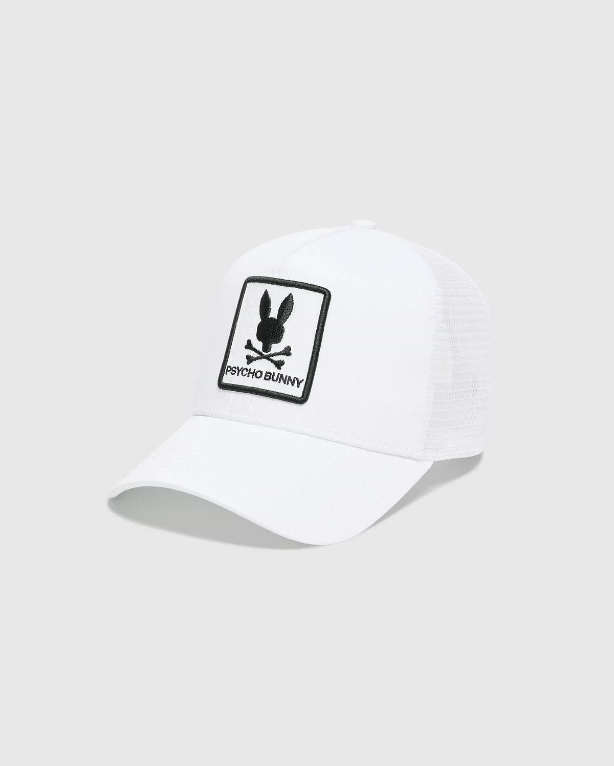 White Mens Adamo Trucker Cap featuring a large, centered black embroidered Psycho Bunny skull and crossbones logo patch with PSYCHO BUNNY text on the solid front panel and a breathable mesh back.
