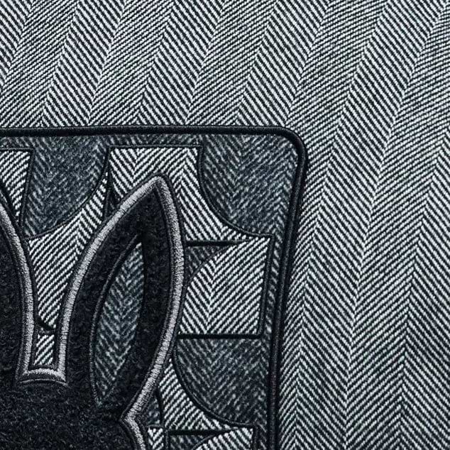 Grey herringbone jacket with black ribbed collar and large embroidered Psycho Bunny skull logo on back, paired with a black Jimmy Baseball Cap featuring a metal logo buckle.