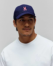 Navy Mens Jimmy Baseball Cap with a pink embroidered Psycho Bunny skull logo, paired with a white relaxed-fit jersey crewneck sweatshirt featuring a small white embroidered Psycho Bunny skull logo on the left chest.