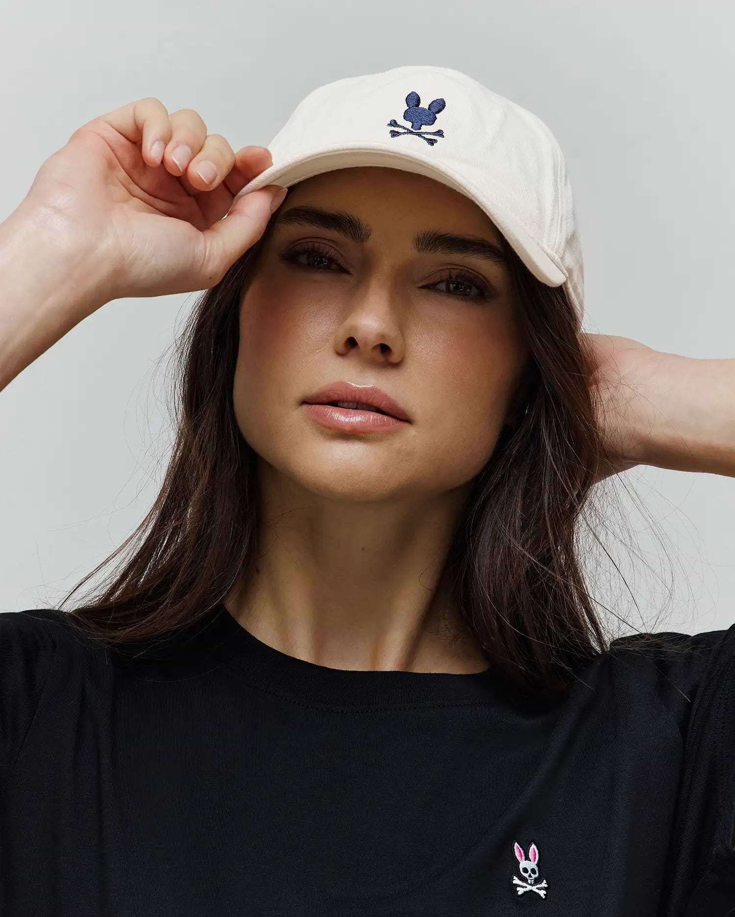 Black classic fit jersey t-shirt with a small embroidered pink and white Psycho Bunny skull logo on the left chest, paired with a Cream Jimmy Baseball Cap featuring a navy embroidered Psycho Bunny logo.