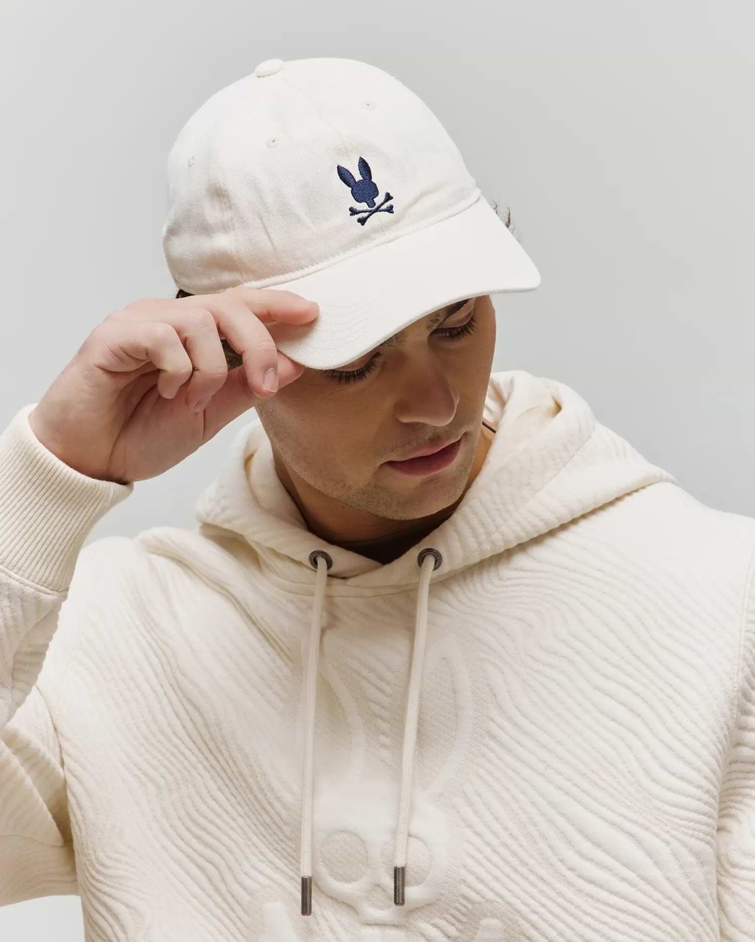 Jimmy Baseball Cap in ecru with a small navy embroidered Psycho Bunny logo, paired with an ecru textured hoodie featuring a wavy pattern and a large tonal skull graphic.