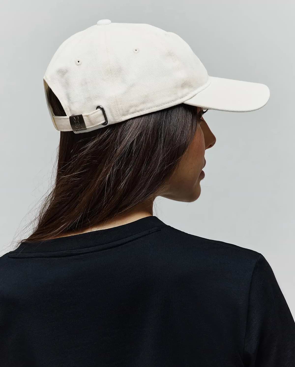 Cream Jimmy Baseball Cap in cotton twill with a dark metal buckle featuring an embossed Psycho Bunny skull logo, paired with a black jersey knit crew neck t-shirt.