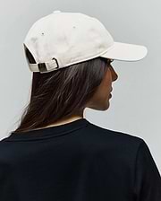 Cream Jimmy Baseball Cap in cotton twill with a dark metal buckle featuring an embossed Psycho Bunny skull logo, paired with a black jersey knit crew neck t-shirt.