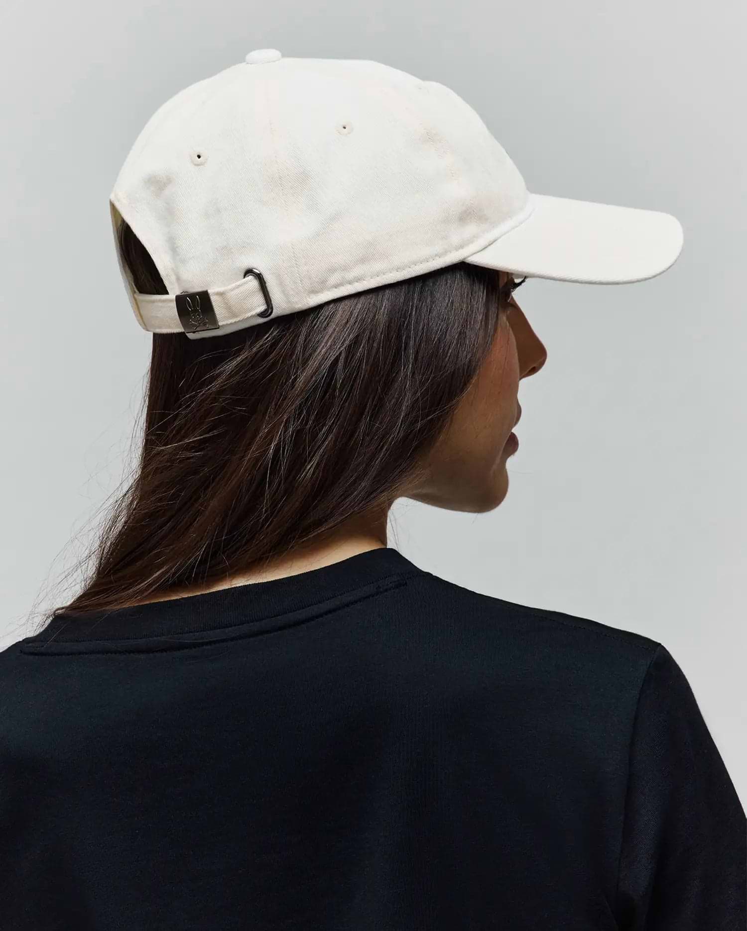 Cream Jimmy Baseball Cap in cotton twill with a dark metal buckle featuring an embossed Psycho Bunny skull logo, paired with a black jersey knit crew neck t-shirt.