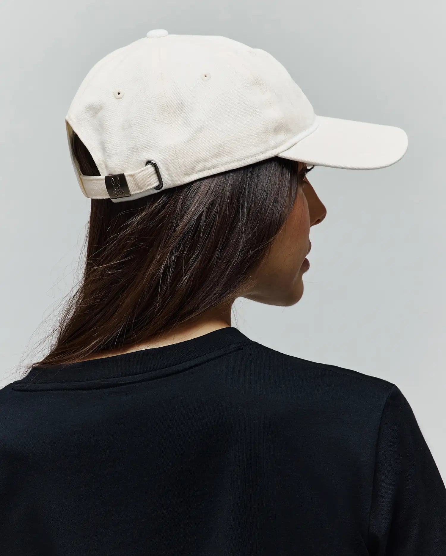 Cream Jimmy Baseball Cap in cotton twill with a dark metal buckle featuring an embossed Psycho Bunny skull logo, paired with a black jersey knit crew neck t-shirt.