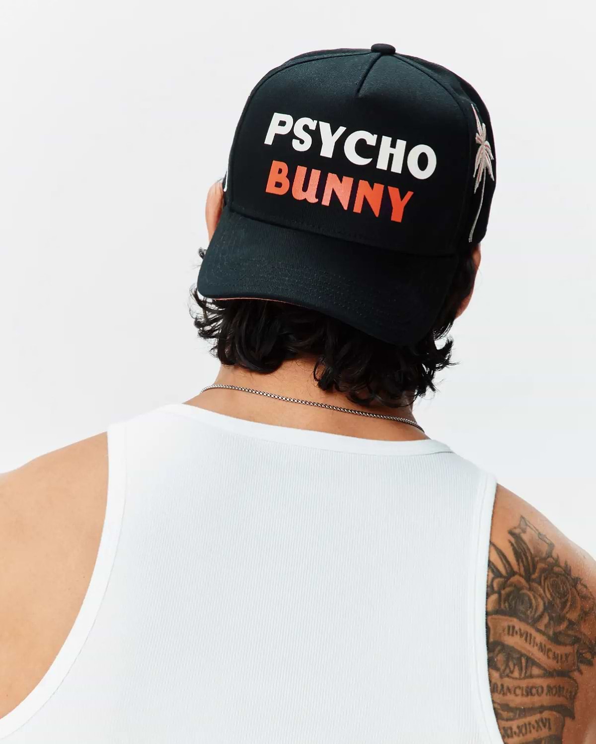 Black Mens Brad Trucker Cap featuring a large white PSYCHO and orange BUNNY text logo on the front, accented by a white palm tree embroidery on the side, paired with a white ribbed tank top.