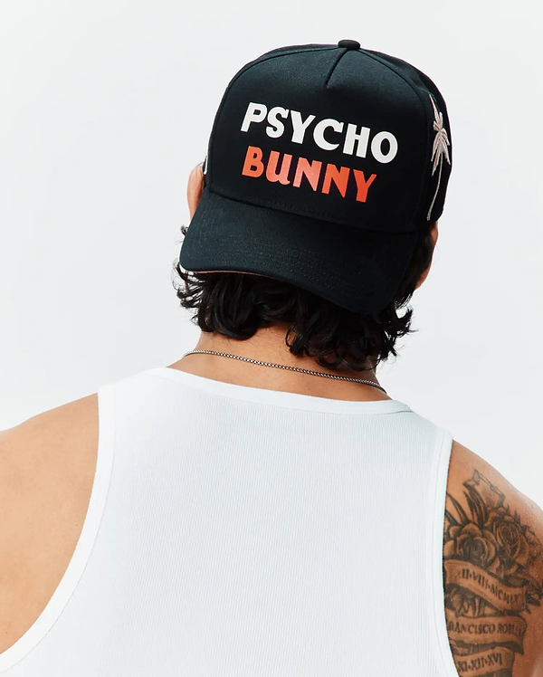 Black Mens Brad Trucker Cap featuring a large white PSYCHO and orange BUNNY text logo on the front, accented by a white palm tree embroidery on the side, paired with a white ribbed tank top.