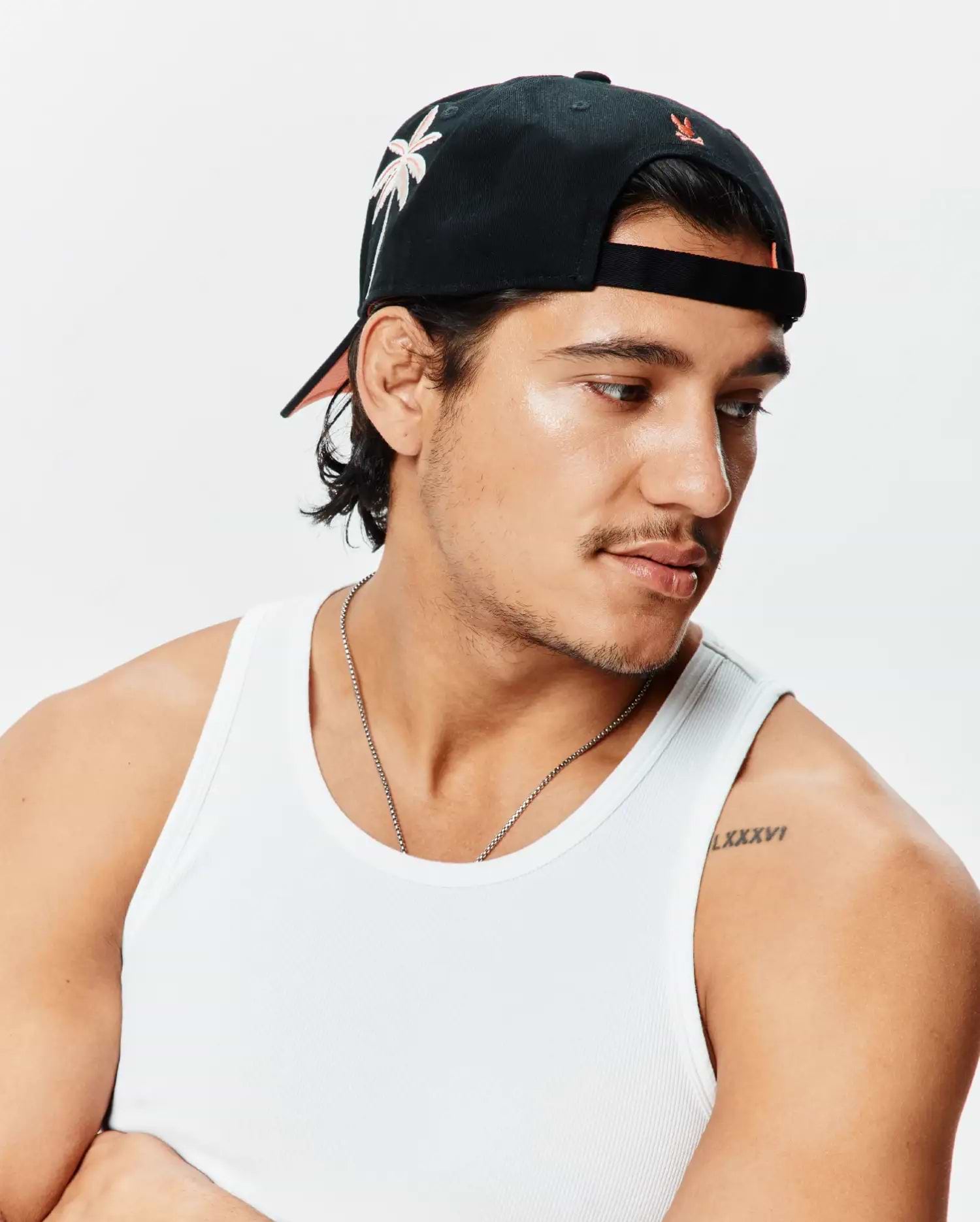 Black Mens Brad Trucker Cap featuring a front light pink embroidered palm tree, small orange Psycho Bunny logo on the back, and peach brim underside, alongside a white ribbed knit classic fit tank top.