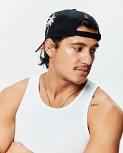 Black Mens Brad Trucker Cap featuring a front light pink embroidered palm tree, small orange Psycho Bunny logo on the back, and peach brim underside, alongside a white ribbed knit classic fit tank top.