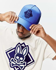 Off-white jersey t-shirt with a large central layered dark blue and white Psycho Bunny skull logo, and a bright blue mesh Mens Tobias Trucker Cap with a dark blue embroidered bunny skull patch.