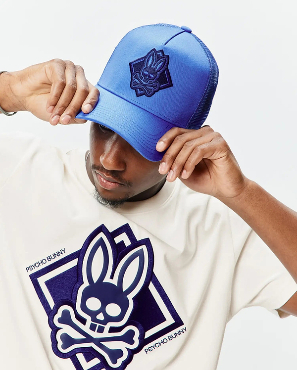 Off-white jersey t-shirt with a large central layered dark blue and white Psycho Bunny skull logo, and a bright blue mesh Mens Tobias Trucker Cap with a dark blue embroidered bunny skull patch.