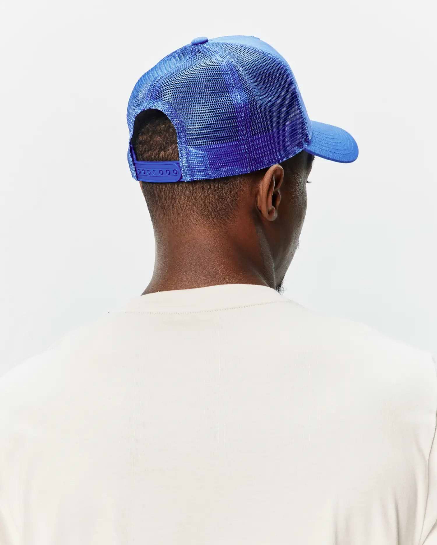 Mens Tobias Trucker Cap in vibrant royal blue, featuring a mesh back, solid front panel, curved brim, and adjustable snap closure, worn with a cream classic fit jersey knit t-shirt.