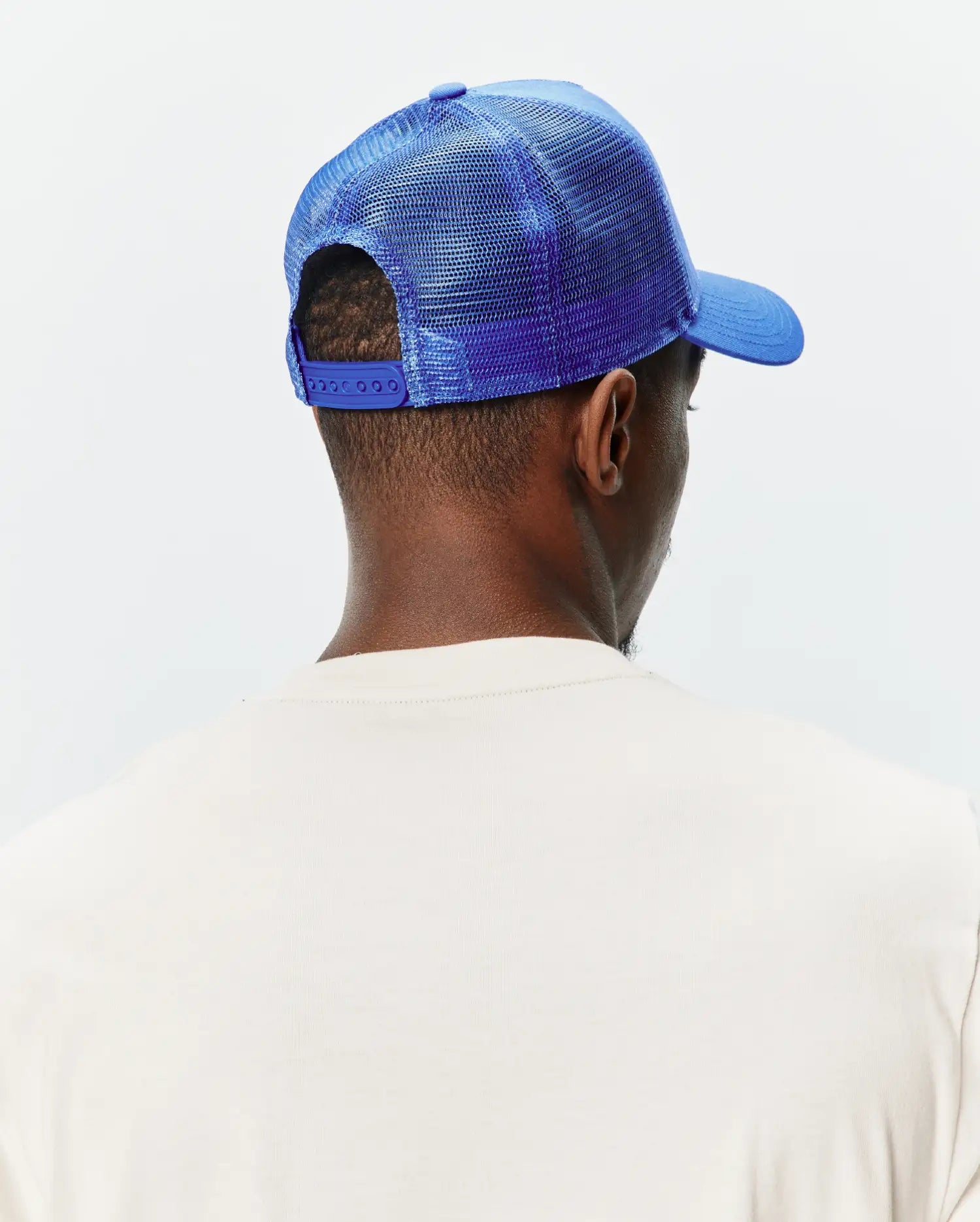 Mens Tobias Trucker Cap in vibrant royal blue, featuring a mesh back, solid front panel, curved brim, and adjustable snap closure, worn with a cream classic fit jersey knit t-shirt.