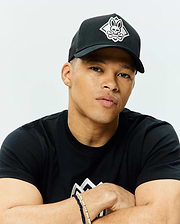 Black Mens Tobias Trucker Cap with a white embroidered Psycho Bunny skull and crossbones logo on the front, featuring a mesh back. Worn with a black classic fit jersey t-shirt displaying a large white geometric chest graphic.