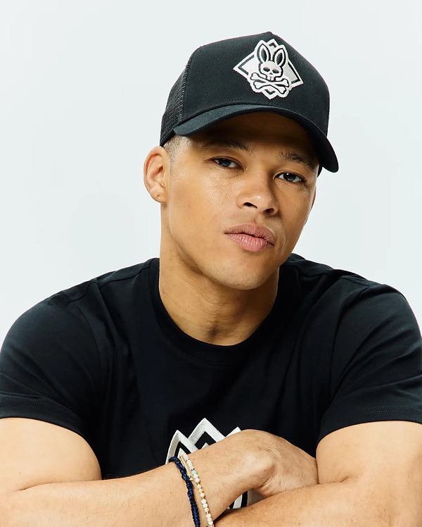 Black Mens Tobias Trucker Cap with a white embroidered Psycho Bunny skull and crossbones logo on the front, featuring a mesh back. Worn with a black classic fit jersey t-shirt displaying a large white geometric chest graphic.