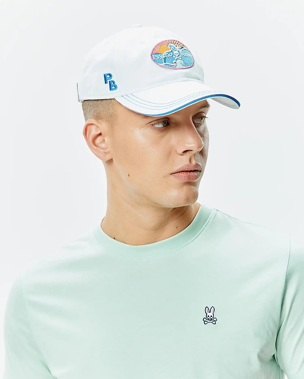 White Mens Ray Baseball Cap with a Psycho Bunny beach patch, blue PB embroidery, and blue brim, alongside a light green jersey crew neck t-shirt with a small embroidered Psycho Bunny skull on the left chest.