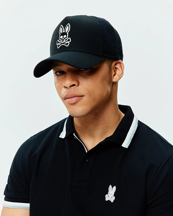 Black pique polo with white/grey striped collar/cuffs and small embroidered Psycho Bunny skull logo on chest, paired with a black Mens Elijah Trucker Cap featuring a large white embroidered Psycho Bunny skull logo on front and mesh back.