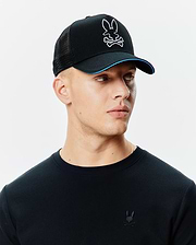 Black Mens Apollo Trucker Cap with mesh back, white embroidered Psycho Bunny skull logo, blue brim accent, and a black pique long-sleeve shirt with a small black embroidered Psycho Bunny logo on the left chest.